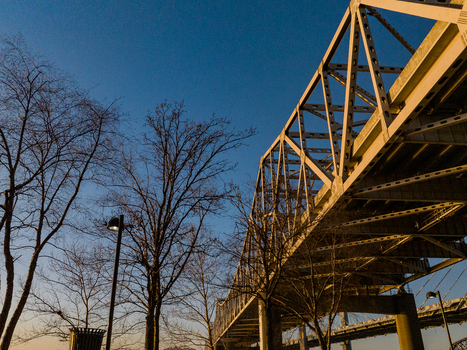 View of the massive steel truss bridge structure under a clear blue sky with bare winter trees in the foreground at dusk in Louisville Kentucky United States. View of the massive steel truss bridge structure under a clear blue sky with bare winter trees in the foreground at dusk in Louisville Kentucky United States.