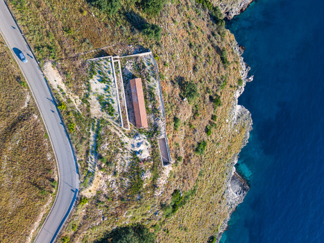 Aerial view of a winding coastal road and a small building with a red roof on a rocky cliff overlooking the blue sea in Porto Palermo Qarku i Vlores Albania. Aerial view of a winding coastal road and a small building with a red roof on a rocky cliff overlooking the blue sea in Porto Palermo Qarku i Vlores Albania.