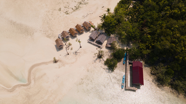 Aerial view of Ransiwor homestay with traditional thatched roof bungalows on a white sand beach wooden pier and turquoise water in Raja Ampat Southwest Papua Indonesia.