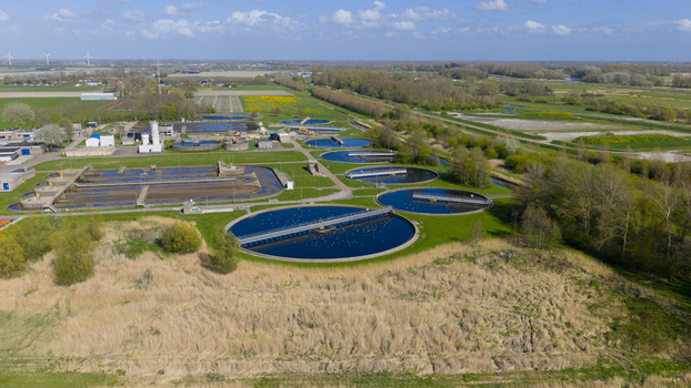 Aerial view of RWZI Geestmerambacht water treatment plant with circular sedimentation tanks and rectangular basins surrounded by green fields and trees under a blue sky. Aerial view of RWZI Geestmerambacht water treatment plant with circular sedimentation tanks and rectangular basins surrounded by green fields and trees under a blue sky.