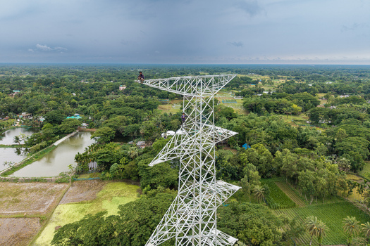 Trishal Upazila Bangladesh 13 May 2026 Aerial view of workers on a tall white electricity pylon surrounded by lush green fields and ponds under an overcast sky. Trishal Upazila Bangladesh 13 May 2026 Aerial view of workers on a tall white electricity pylon surrounded by lush green fields and ponds under an overcast sky.