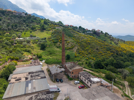 Kruje Albania 12 June 2026 Aerial view of an old industrial facility with a tall brick chimney stack surrounded by lush green hills and olive groves.