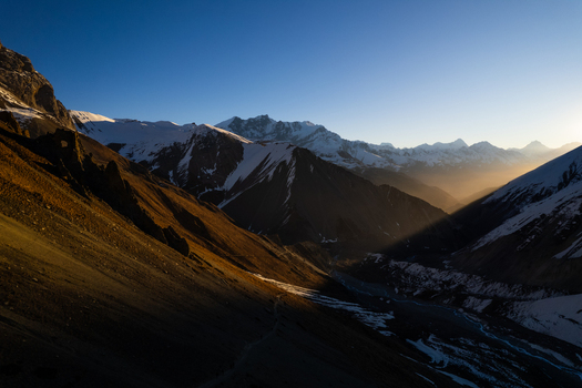 Aerial view of the Himalayan mountain range featuring snow capped peaks deep valleys and dramatic golden hour light casting long shadows across the rugged terrain in Nepal.