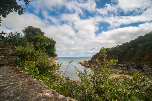 View of the Fowey River estuary with boats rocky coastline lush greenery and a stone wall in the foreground under a cloudy blue sky in Fowey Cornwall United Kingdom. View of the Fowey River estuary with boats rocky coastline lush greenery and a stone wall in the foreground under a cloudy blue sky in Fowey Cornwall United Kingdom.