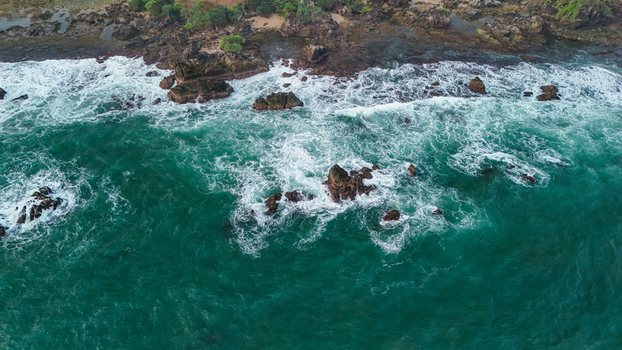 Aerial view of a rocky coastline with turquoise waves crashing against dark volcanic rocks and lush green vegetation along the shore in Indonesia. Aerial view of a rocky coastline with turquoise waves crashing against dark volcanic rocks and lush green vegetation along the shore in Indonesia.