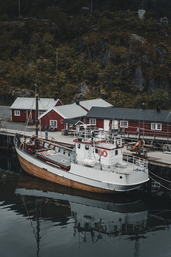Nusfjord Norway 07 September 2022 View of a fishing boat docked at the pier in the historic village of Nusfjord with red wooden cabins and rocky hills. Nusfjord Norway 07 September 2022 View of a fishing boat docked at the pier in the historic village of Nusfjord with red wooden cabins and rocky hills.
