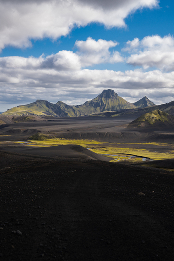 View of vast dark volcanic plains meet moss covered mountains under a bright sky streaked with clouds Skaftárhreppur Iceland. View of vast dark volcanic plains meet moss covered mountains under a bright sky streaked with clouds Skaftárhreppur Iceland.