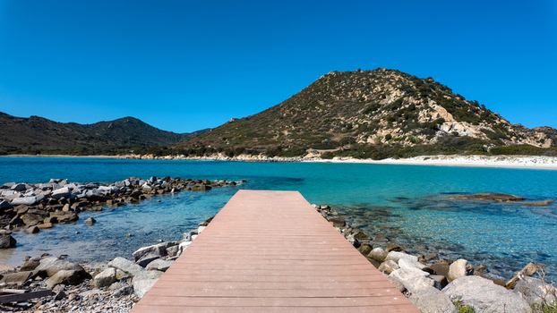 View of Punta Molentis Beach with a wooden pier leading into turquoise water white sand and rugged hills under a clear blue sky Villasimius Sardinia Italy. View of Punta Molentis Beach with a wooden pier leading into turquoise water white sand and rugged hills under a clear blue sky Villasimius Sardinia Italy.