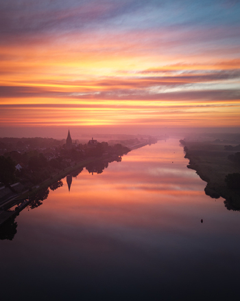 Aerial view of the Meuse river reflecting a vibrant sunrise sky with the historic town silhouette and church spire under a morning mist Kessel Limburg Netherlands.