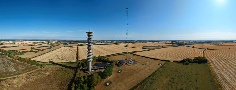 Aerial view of the Morborne Hill Transmitter a tall concrete tower and a slender mast standing amidst golden harvested fields under a clear blue sky in Morborne England United Kingdom. Aerial view of the Morborne Hill Transmitter a tall concrete tower and a slender mast standing amidst golden harvested fields under a clear blue sky in Morborne England United Kingdom.