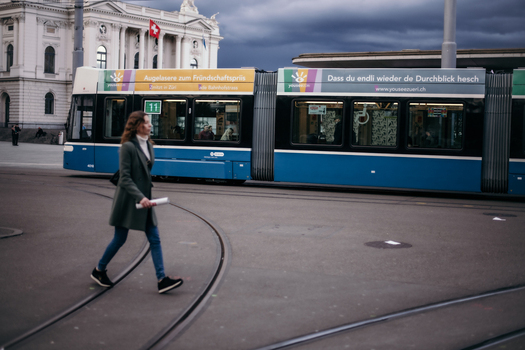 Zurich Switzerland 02 April 2023 View of a woman crossing the tram tracks as a blue and white tramcar glides by reflecting the overcast sky. Zurich Switzerland 02 April 2023 View of a woman crossing the tram tracks as a blue and white tramcar glides by reflecting the overcast sky.
