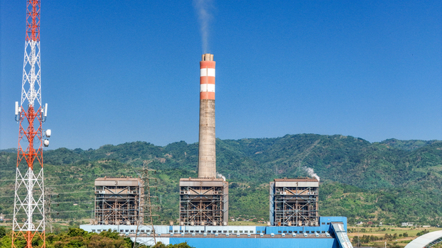 Aerial view of a power plant with a tall chimney emitting smoke industrial buildings and a communication tower set against a backdrop of lush green mountains in Indonesia.