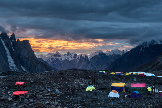 View of Concordia Campsite nestled among rugged Karakoram peaks under a dramatic sunset sky with colorful tents on the rocky glacier terrain in Concordia Gilgit Baltistan Pakistan.