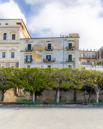 View of aged buildings stand above a line of vibrant green trees their leaves contrasting with the peeling paint and blue sky Ortigia Sicily Italy.