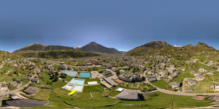 Aerial view of the town of Naters featuring outdoor swimming pools residential buildings and lush green fields surrounded by the Swiss Alps under a clear blue sky Naters Valais Switzerland.