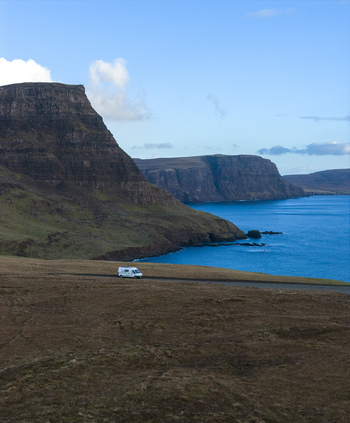 Aerial view of a white campervan driving along the coastal road near the steep cliffs of Neist Point overlooking the blue sea in Isle of Skye Scotland United Kingdom. Aerial view of a white campervan driving along the coastal road near the steep cliffs of Neist Point overlooking the blue sea in Isle of Skye Scotland United Kingdom.