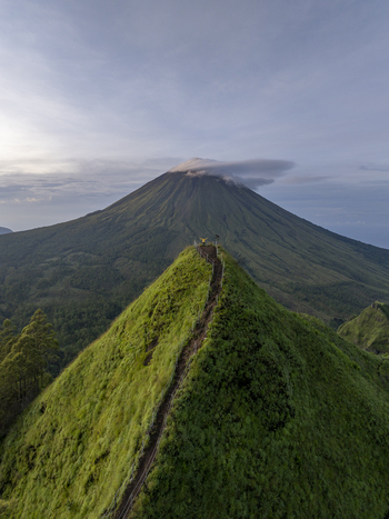 Aerial view of Mount Inerie with a person standing on a lush green mountain ridge under a soft sky in Inerie Flores Indonesia. Aerial view of Mount Inerie with a person standing on a lush green mountain ridge under a soft sky in Inerie Flores Indonesia.