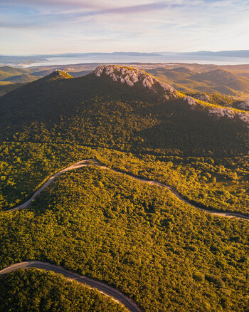 Aerial view of a winding mountain road snaking through lush green forests and rocky peaks under a soft golden sunset light with the Adriatic Sea in the distance Rijeka Primorje Gorski Kotar Croatia. Aerial view of a winding mountain road snaking through lush green forests and rocky peaks under a soft golden sunset light with the Adriatic Sea in the distance Rijeka Primorje Gorski Kotar Croatia.