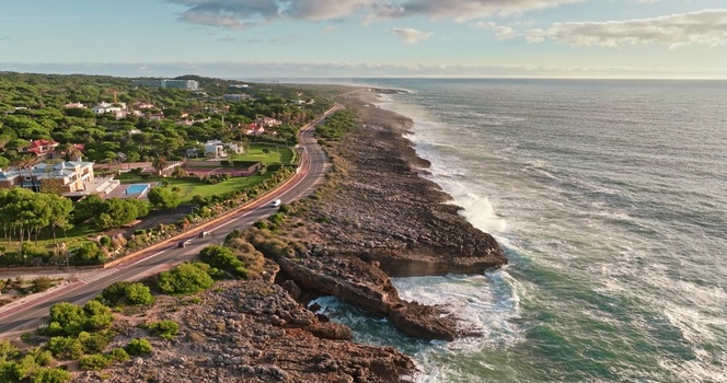 Aerial view of a coastal road winding along rocky cliffs and the Atlantic Ocean bordered by lush green trees and residential villas in Lisbon Lisbon Portugal. Aerial view of a coastal road winding along rocky cliffs and the Atlantic Ocean bordered by lush green trees and residential villas in Lisbon Lisbon Portugal.