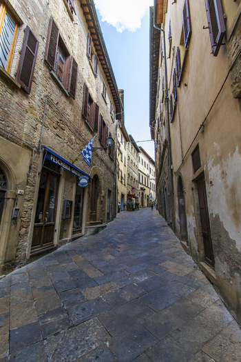 View of a narrow street paved with stone under a blue sky flanked by aged buildings with weathered walls and shuttered windows Volterra Tuscany Italy. View of a narrow street paved with stone under a blue sky flanked by aged buildings with weathered walls and shuttered windows Volterra Tuscany Italy.