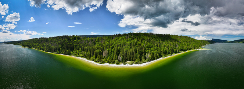 Aerial view of the vibrant green forest meeting the tranquil Lac de Joux under a sky of shifting clouds Le Lieu Vaud Switzerland.