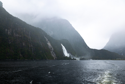View of cascading waterfalls plunge from verdant mist shrouded cliffs into the dark rippled waters below creating an ethereal scene Milford Sound Southland Region New Zealand.
