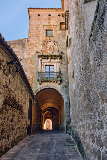 Plasencia Spain 03 December 2025 View of a narrow stone alleyway passing under a historic archway with a decorative balcony and coat of arms in the old town. Plasencia Spain 03 December 2025 View of a narrow stone alleyway passing under a historic archway with a decorative balcony and coat of arms in the old town.