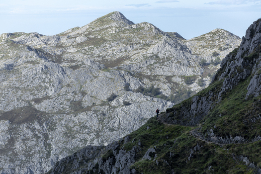 View of a solitary figure traversing a steep grassy path amidst the rugged grey white peaks under a soft blue sky in Picos de Europa Spain. View of a solitary figure traversing a steep grassy path amidst the rugged grey white peaks under a soft blue sky in Picos de Europa Spain.