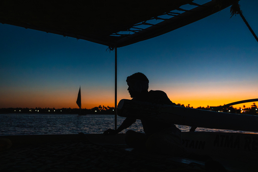 View of the silhouette of a man on a traditional boat during sunset on the Nile River with a felucca in the distance Aswan Aswan Governorate Egypt. View of the silhouette of a man on a traditional boat during sunset on the Nile River with a felucca in the distance Aswan Aswan Governorate Egypt.
