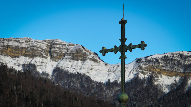 Aerial view of the metal cross of Eglise Saint Pierre with snow capped mountains and a clear blue sky in the background Gex Auvergne Rhone Alpes France.