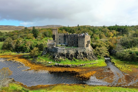 Aerial view of Dunvegan Castle perched on a rocky outcrop above a tidal inlet surrounded by dense woodland and rolling hills in Isle of Skye Scotland United Kingdom. Aerial view of Dunvegan Castle perched on a rocky outcrop above a tidal inlet surrounded by dense woodland and rolling hills in Isle of Skye Scotland United Kingdom.