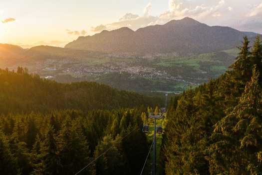 Aerial view of Fiemme Valley with a cable car gliding over dense pine forests during golden hour in Trentino Alto Adige Italy.