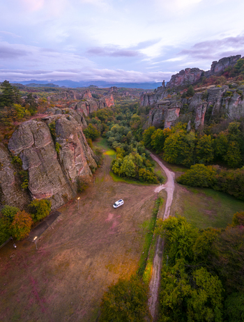 Aerial view of red sandstone rock formations of Belogradchik with a white camper van parked on a dirt path surrounded by lush autumn forests in Belogradchik Vidin Bulgaria. Aerial view of red sandstone rock formations of Belogradchik with a white camper van parked on a dirt path surrounded by lush autumn forests in Belogradchik Vidin Bulgaria.