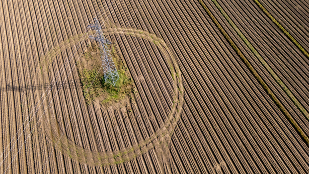 Aerial view of a high voltage electricity pylon standing in the center of a large plowed agricultural field with circular tracks and parallel furrows in Poland.