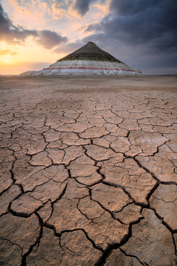 View of the Sherkala mountain rising from the cracked arid desert floor under a dramatic sunset sky in Mangistau Mangystau Region Kazakhstan. View of the Sherkala mountain rising from the cracked arid desert floor under a dramatic sunset sky in Mangistau Mangystau Region Kazakhstan.