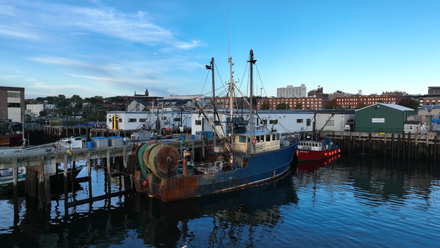 Aerial view of a blue fishing boat docked at the historic Portland waterfront and fishing piers with calm water and urban buildings in Portland Maine United States.