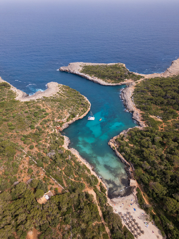 Aerial view of Cala Sa Nau cove featuring turquoise water rocky cliffs with pine trees and a white boat anchored in the bay in Felanitx Balearic Islands Spain. Aerial view of Cala Sa Nau cove featuring turquoise water rocky cliffs with pine trees and a white boat anchored in the bay in Felanitx Balearic Islands Spain.