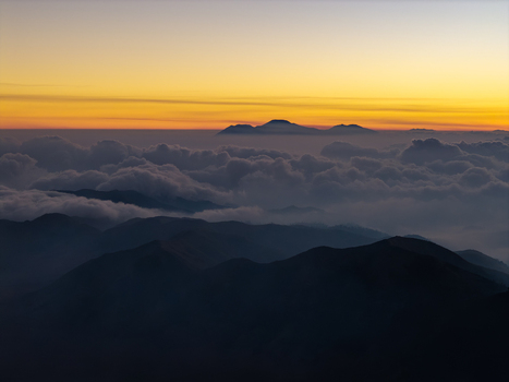 Aerial view of Mount Bromo at sunset with golden light illuminating the horizon above a thick sea of clouds and dark mountain silhouettes in Tosari East Java Indonesia. Aerial view of Mount Bromo at sunset with golden light illuminating the horizon above a thick sea of clouds and dark mountain silhouettes in Tosari East Java Indonesia.