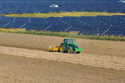 Aerial view of a green tractor plowing a field in front of a large solar farm with rows of photovoltaic panels in Poland.