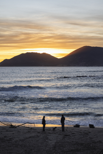 View of Venere Azzurra beach with silhouettes of two people and a dog at sunset against a backdrop of mountains and waves in Lerici Liguria Italy.