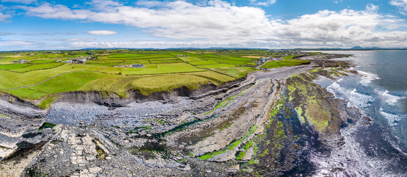 Aerial view of the rugged coastline and rocky shore featuring green fields dramatic cliffs and the Atlantic Ocean under a blue sky Enniscrone County Sligo Ireland. Aerial view of the rugged coastline and rocky shore featuring green fields dramatic cliffs and the Atlantic Ocean under a blue sky Enniscrone County Sligo Ireland.