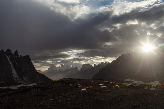 View of Concordia Campsite nestled among dramatic mountain peaks under a stormy sky with sunburst featuring colorful tents on the rocky glacier terrain in Concordia Gilgit Baltistan Pakistan.