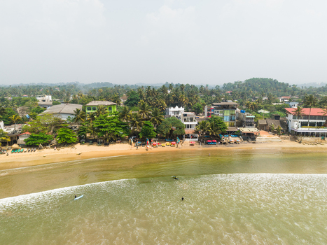 Aerial view of Dewata Beach Road with surfers in the ocean coastal buildings and lush palm trees Galle Southern Province Sri Lanka.