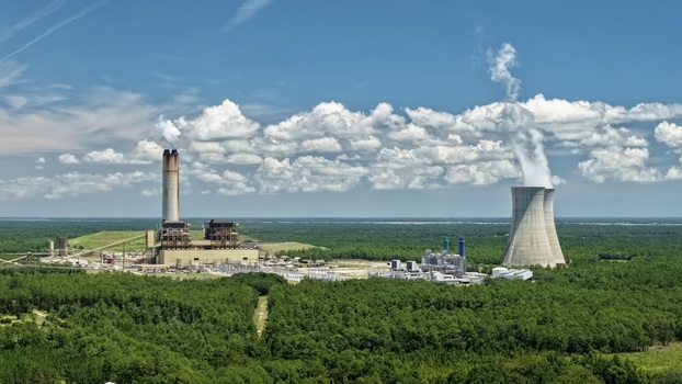 Aerial view of the Seminole Electric power plant featuring a tall smokestack with steam a large cooling tower and industrial infrastructure surrounded by dense green forest in Palatka Florida United States.