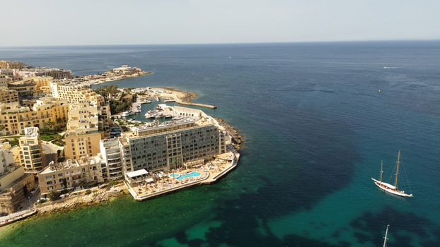 Aerial view of a coastal hotel resort with a swimming pool sun loungers and a sailboat anchored in the clear turquoise Mediterranean sea in Malta. Aerial view of a coastal hotel resort with a swimming pool sun loungers and a sailboat anchored in the clear turquoise Mediterranean sea in Malta.