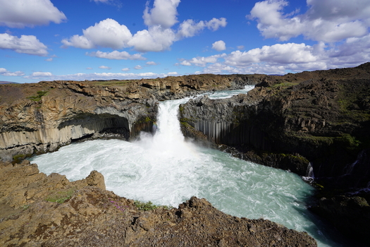 View of the powerful Aldeyjarfoss waterfall cascading over basalt columns into a turquoise pool under a bright blue sky with fluffy clouds in Iceland. View of the powerful Aldeyjarfoss waterfall cascading over basalt columns into a turquoise pool under a bright blue sky with fluffy clouds in Iceland.