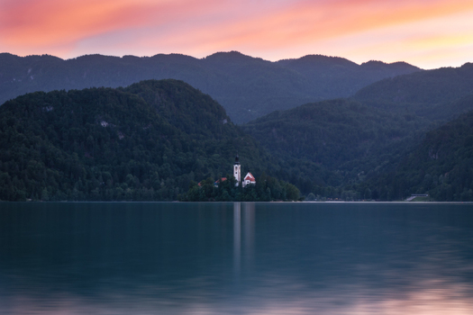 View of the Pilgrimage Church of the Assumption of Mary on Lake Bled island surrounded by forested mountains under a sunset sky in Bled Radovljica Slovenia. View of the Pilgrimage Church of the Assumption of Mary on Lake Bled island surrounded by forested mountains under a sunset sky in Bled Radovljica Slovenia.