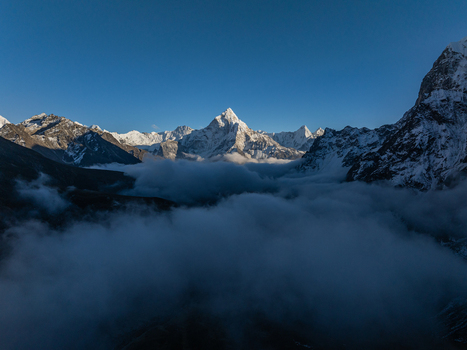 Aerial view of snow capped Himalayan peaks rising above a thick layer of clouds under a clear blue sky in Nepal.