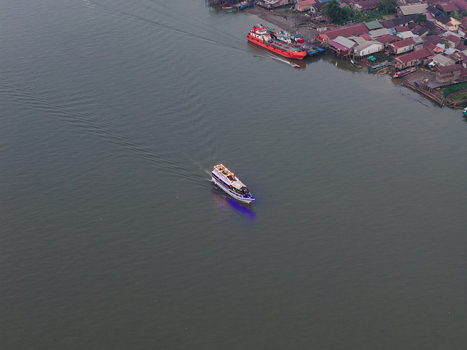 Aerial view of a passenger boat cruising along the Mahakam River past a riverside settlement with stilt houses and a docked cargo vessel in Samarinda East Kalimantan Indonesia. Aerial view of a passenger boat cruising along the Mahakam River past a riverside settlement with stilt houses and a docked cargo vessel in Samarinda East Kalimantan Indonesia.