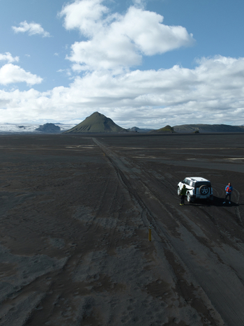 Aerial view of Maelifell volcano in a black sand desert with a white off road vehicle and two people standing nearby under a blue sky in Kirkjubaejarklaustur Skaftarhreppur Iceland. Aerial view of Maelifell volcano in a black sand desert with a white off road vehicle and two people standing nearby under a blue sky in Kirkjubaejarklaustur Skaftarhreppur Iceland.